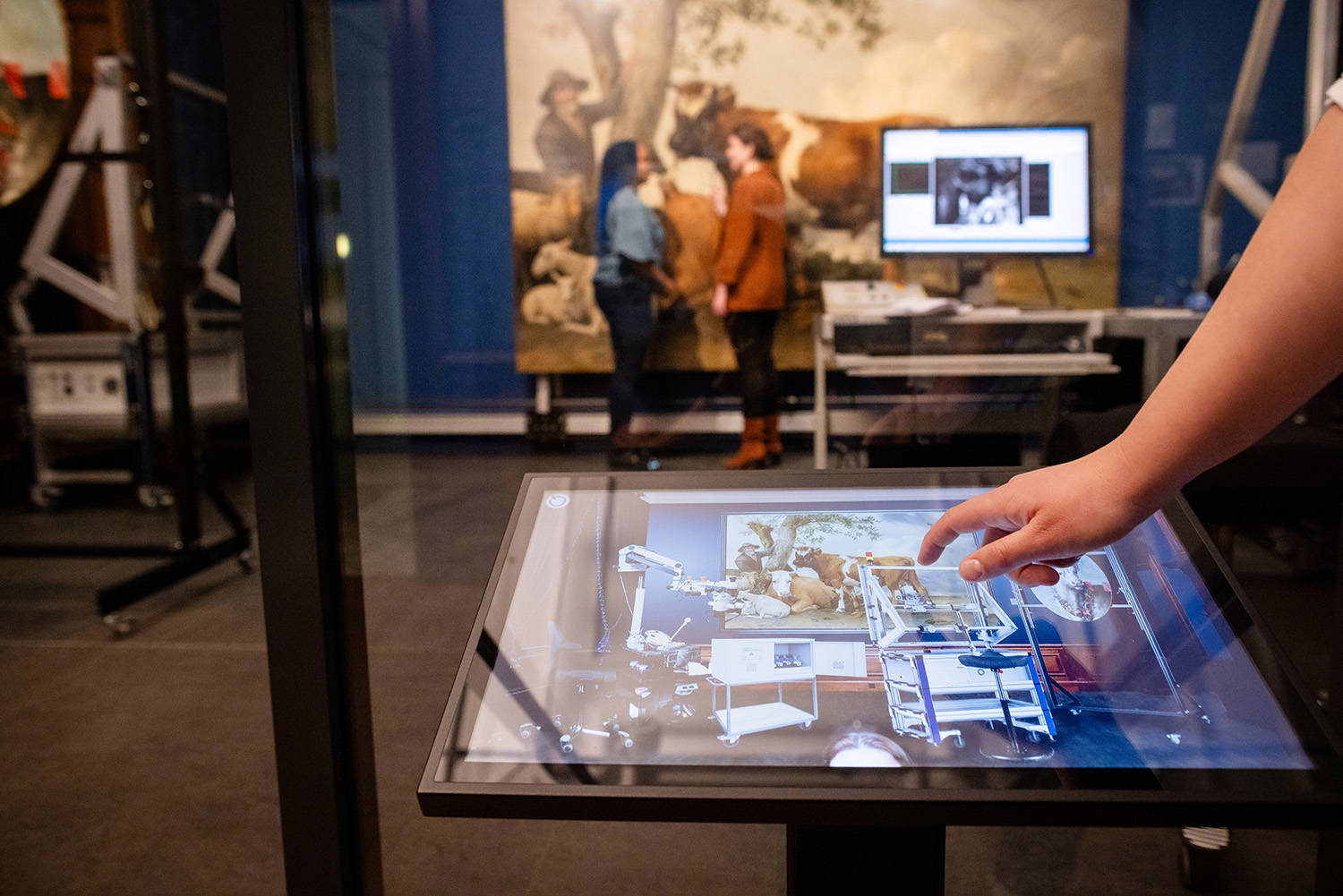 Een hand gaat over het scherm bij de restauratie van de Stier. Op de achtergrond het schilderij De Stier van Paulus Potter en de twee conservatoren.