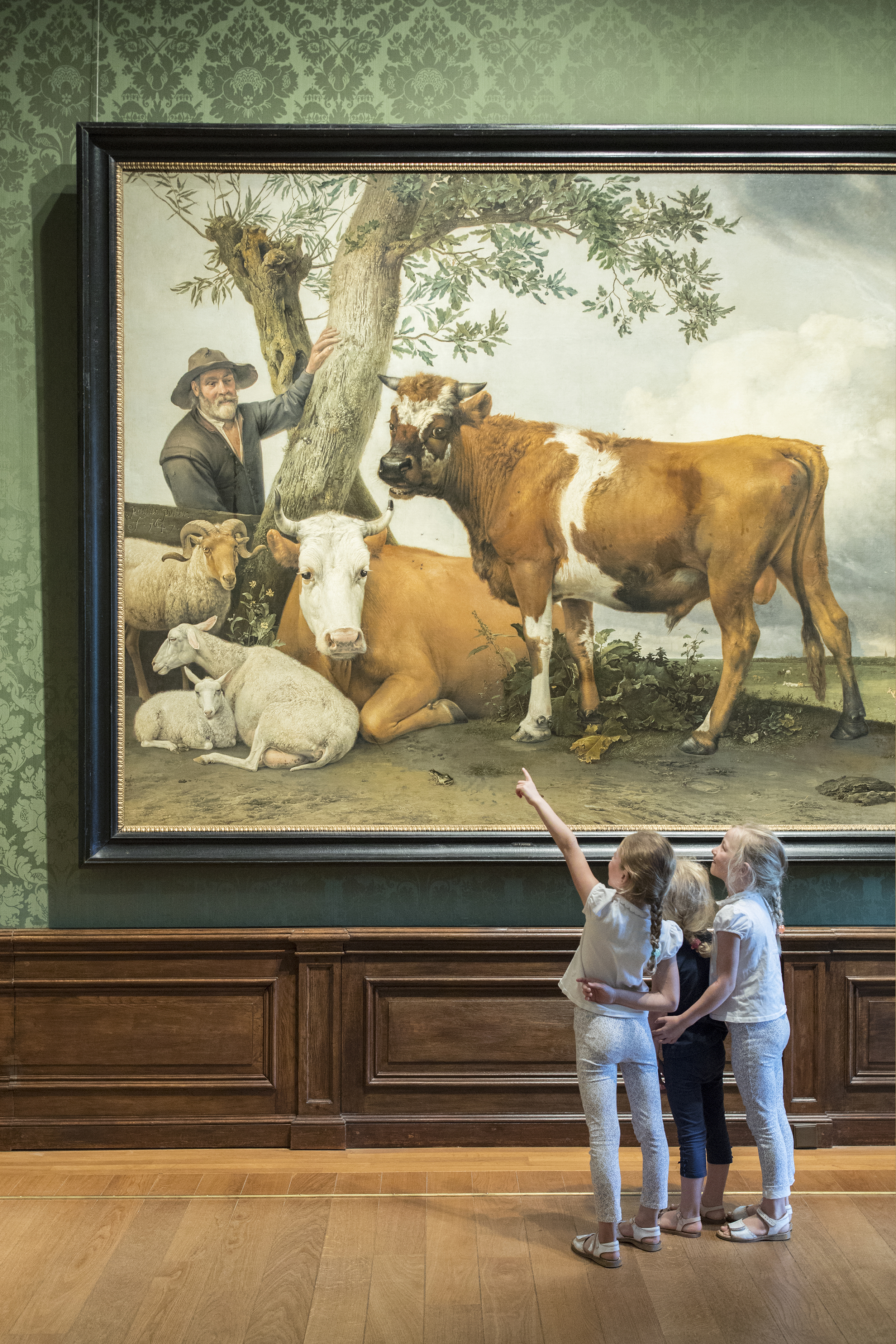 Drie kinderen staan en wijzen bij het beroemde schilderij 'De stier' van Paulus Potter.
