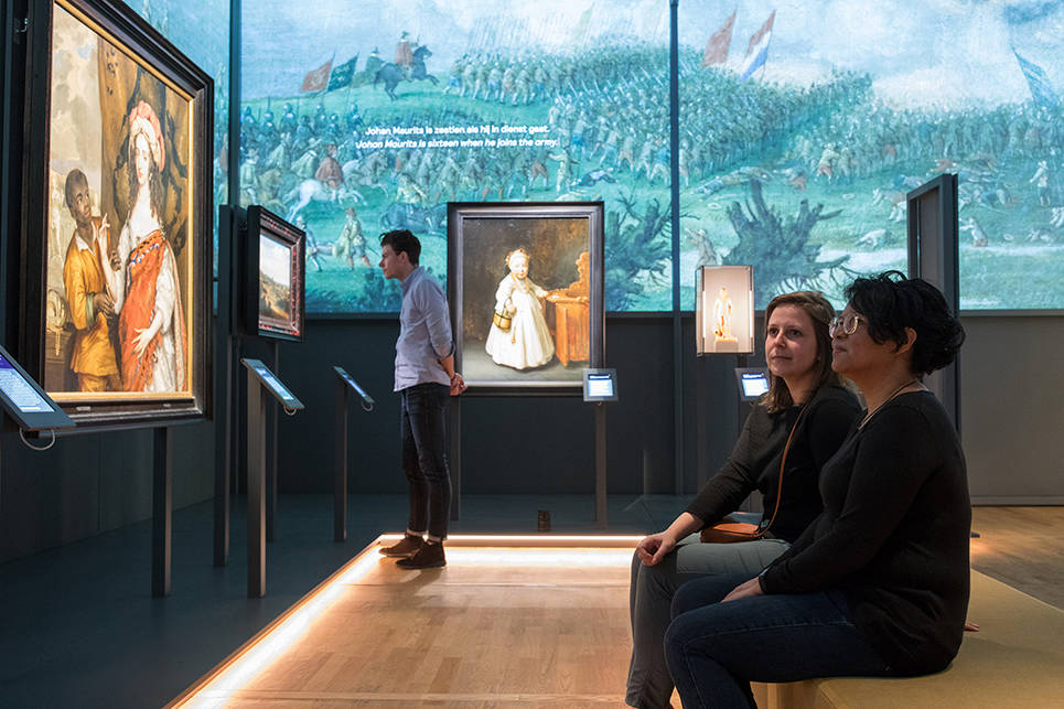 Een overzichtsfoto van de tentoonstelling Bewogen Beeld. Vooraan zitten twee vrouwen, ze kijken naar een schilderij.