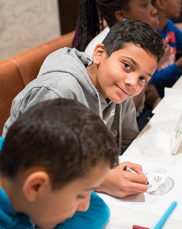 Een kind tekent tijdens een kinderactiviteit in het Mauritshuis in Den Haag. Hij lacht naar de camera.