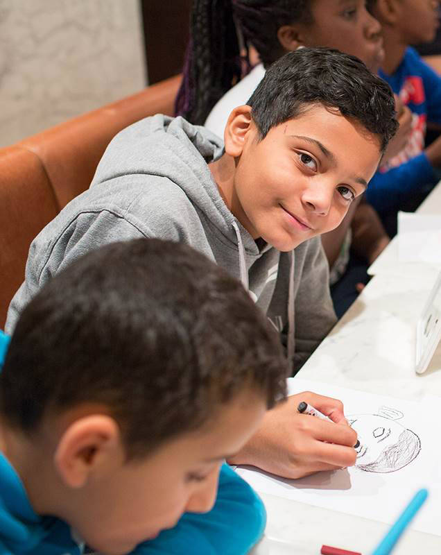Een kind tekent tijdens een kinderactiviteit in het Mauritshuis in Den Haag. Hij lacht naar de camera.