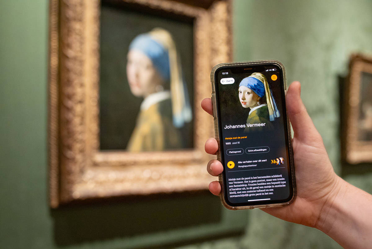 Een hand met een telefoon toont de app van het Mauritshuis. Op de achtergrond hangt Meisje met de parel van Johannes Vermeer.