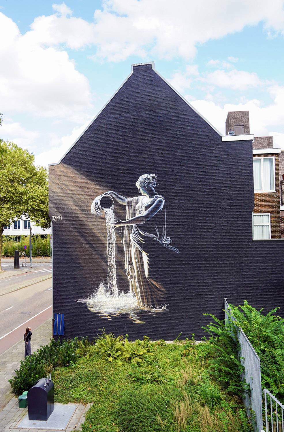 Een vrouw kijkt naar de mural van Ozmo, geinspireerd door het Romeins Museum. Een vrouw in Romeinse klederdracht giet water uit een kruik, geschilderd in wit op zwart.