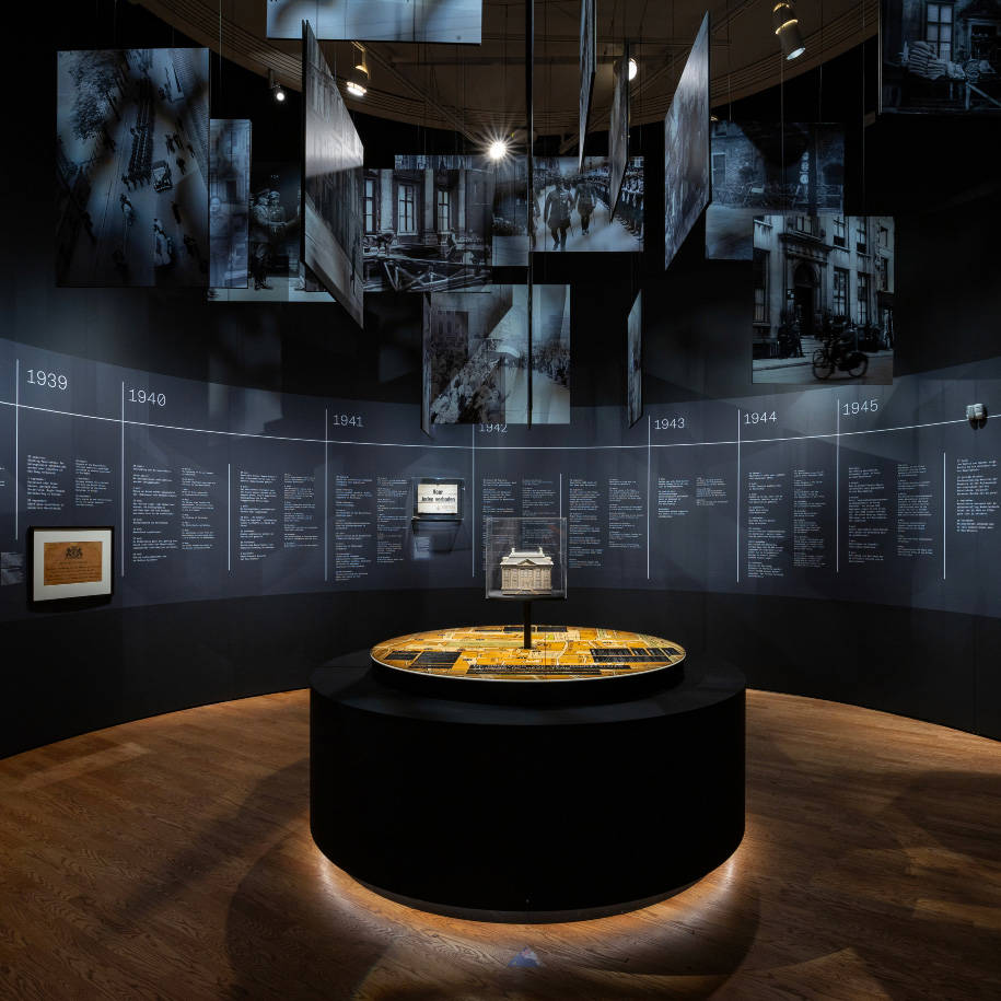 De tentoonstellingszaal van Huis in de Storm. Er is een tijdlijn, bovenaan hangen foto's uit de oorlog als wolken en in het midden een replica van het Mauritshuis.