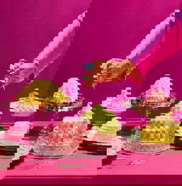 Een tafel voor kleurrijke toetjes tegen een roze achtergrond. Een hand staat op het punt een prikkertje in één van de toetjes te steken.