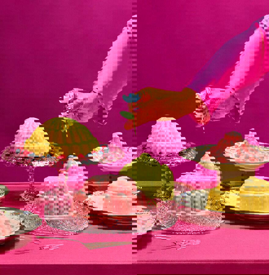 Een tafel voor kleurrijke toetjes tegen een roze achtergrond. Een hand staat op het punt een prikkertje in één van de toetjes te steken.